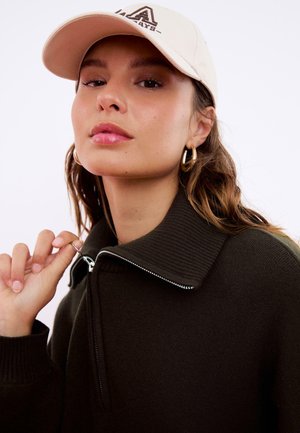 Sweater zippé vert foncé avec col côtelé, associé à une casquette beige ornée d'un logo. Le modèle tient la tirette du zip d'une main.