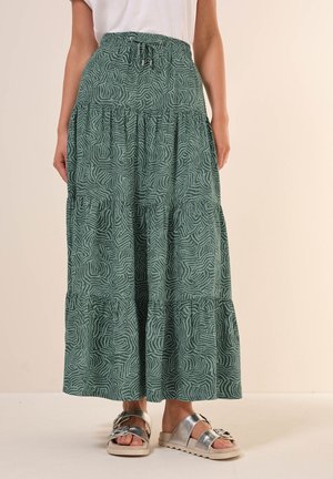 Femme portant une jupe longue verte à motifs à volants avec une taille élastique et des sandales argentées à bout ouvert avec des brides épaisses.