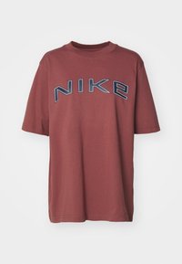 Rostfärgad kortärmad T-shirt med en marinblå och vit konturerad "NIKE"-logotyp över bröstet på en neutral vit bakgrund.
