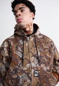 Bruine camouflage zip-up hoodie met een gladde textuur, zwarte trekkoorden en een geborduurd logo op de voorzak.