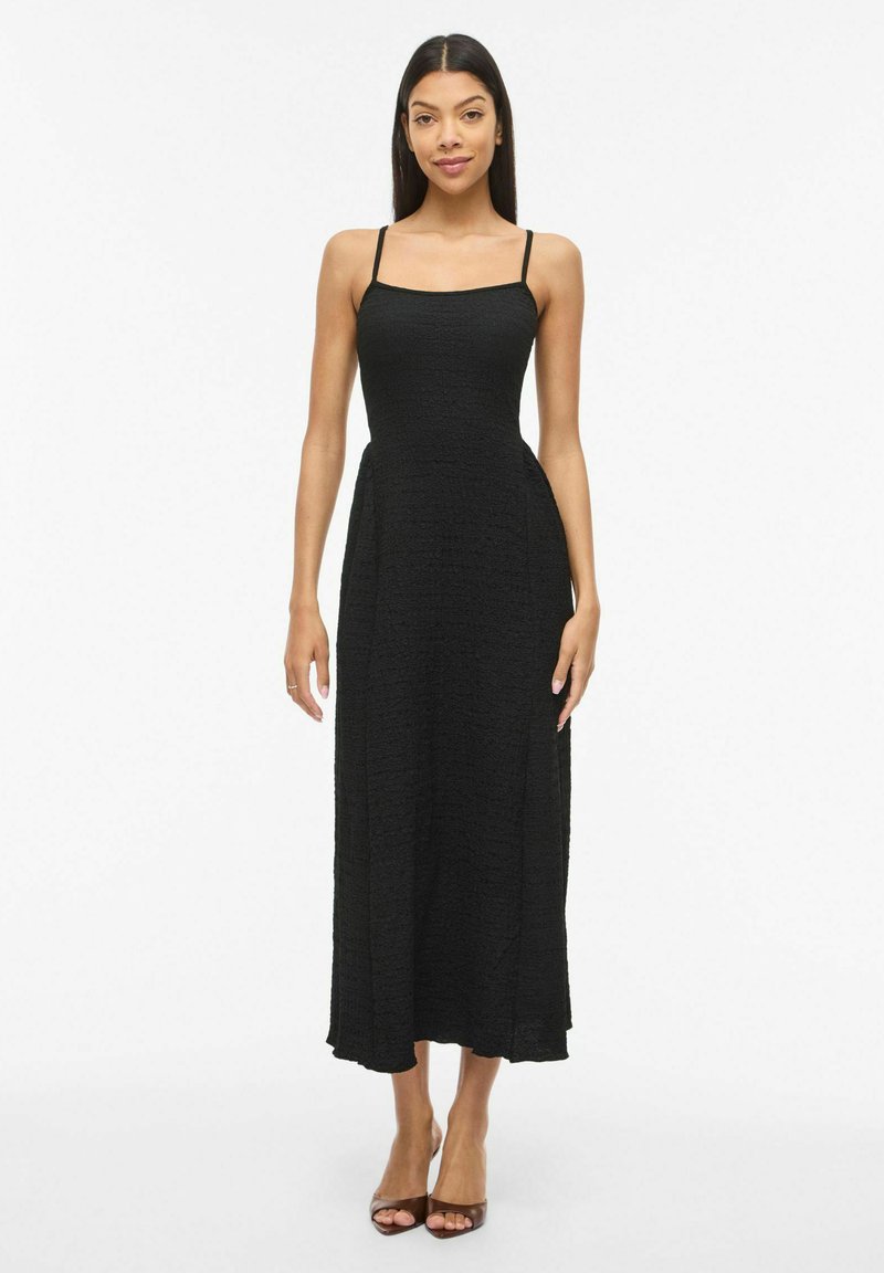 Femme portant une robe midi noire sans manches avec fines bretelles et des escarpins à bout ouvert, debout devant un fond blanc.
