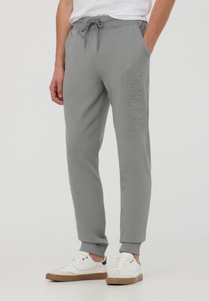 Pantaloni felpati grigi con vita elasticizzata, cordoncino regolabile, tasche laterali e dettaglio in rilievo sul lato sinistro. Tessuto dalla consistenza morbida.