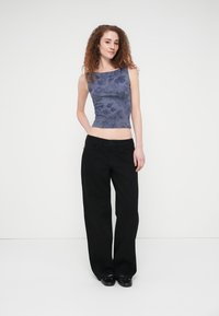 Μπλε floral crop top, χωρίς μανίκια, εφαρμοστή σχεδίαση. Συνδυάζεται με άνετο μαύρο παντελόνι. Το μοντέλο στέκεται μπροστά σε λευκό, απλό φόντο.
