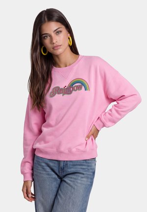 Giovane donna con lunghi capelli castani, che indossa orecchini a cerchio gialli, felpa rosa con "Rainbow" e una grafica arcobaleno, e jeans blu, posando con la mano sul fianco.