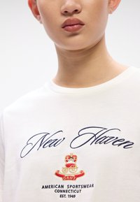 White cotton t-shirt featuring blue embroidered text "New Haven" and a colorful coat of arms logo with details below. Round neckline.