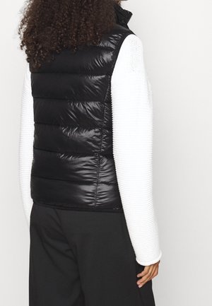 Personne aux cheveux bouclés portant une veste matelassée noire sur un haut blanc texturé à manches longues et un pantalon noir, dos à la caméra.