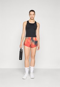 Svart atletisk tank top, flerfargede shorts med gradientmønster, hvite joggesko, og en svart vannflaske i hånden.