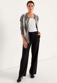 Vrouw staat met één hand in de zak, draagt een grijze gebreide cardigan, een wit topje, zwarte wijde broek en zwarte sneakers.
