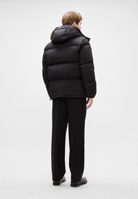 Crna puf jakna s kapuljačom, s kvadratnim dizajnom i elastičnom fudbom. Nosi se s crnim širokim hlačama i ravnim cipelama.