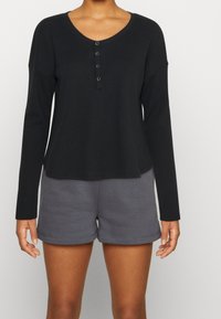 Person klädd i svart långärmad waffle stickad henley-top och mörkgrå avslappnade shorts, stående mot en ljus och enhetlig bakgrund.