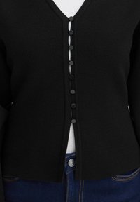 Gros plan d’un cardigan noir boutonné porté sur un top blanc et associé à un jean en denim bleu.