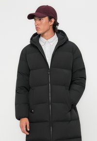 Zwarte pufferjas met een capuchon, voorzien van een rits aan de voorkant, een gequilt ontwerp en zijzakken. Draagend over een gestreept overhemd met knopen en een bordeauxrode pet.