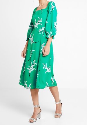 Grøn blomsterkjole med puffede ærmer, firkantet udskæring og hvide blomsterprint. Kombineret med sølvfarvede sandaler med blokhæle.