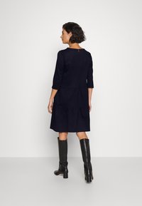 Dunkelblaues Kleid mit Dreiviertelärmeln, gestuftem Rock, rundem Ausschnitt und Schlüssellochdetail im Rücken, kombiniert mit knielangen schwarzen Stiefeln.