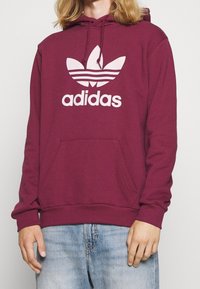 Mörkröd huvtröja i mjukt tyg, med en stor vit Adidas-logga och den klassiska tre-randiga designen. Inkluderar en känguruficka och huva.