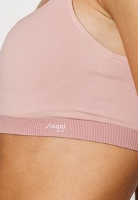 Svijetloružičasti sportski grudnjak izrađen od glatkog materijala s rebrastim elastičnim pojasom. Na pojasu se suptilno ispisuje logo brenda u bijeloj boji.