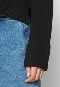 Mano appoggiata sul fianco, indossando un maglione nero a coste con maniche lunghe e jeans di denim blu.