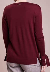 Maglione bordeaux con scollatura tonda, maniche lunghe con dettagli a fiocco e spacco laterale all'orlo. Tessuto morbido, polsini a coste.
