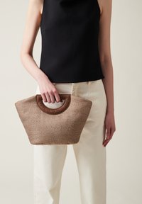 Borsa marrone texturizzata con un manico rotondo marrone, dotata di un'accoppiatura con logo e un ampio top aperto. Tenuta da una persona, abbinata a pantaloni chiari.
