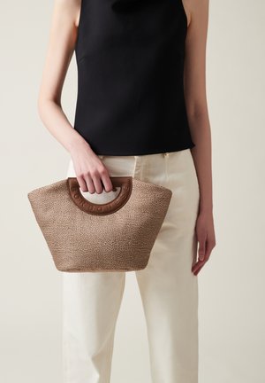 Borsa marrone texturizzata con un manico rotondo marrone, dotata di un'accoppiatura con logo e un ampio top aperto. Tenuta da una persona, abbinata a pantaloni chiari.
