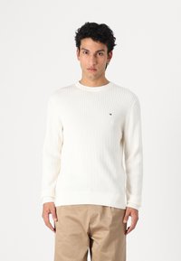 Suéter blanco de punto con un patrón texturizado de diamantes, puños acanalados y cuello redondo, combinado con pantalones beiges. Pequeño logotipo en el pecho.