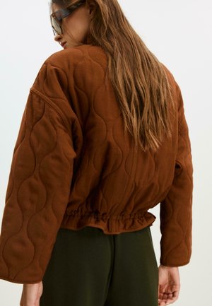 Braune gesteppte Jacke mit tailliertem Schnitt, übergroßen Ärmeln und strukturierten Wellenmustern auf dem Stoff, kombiniert mit dunkelgrünen Hosen.
