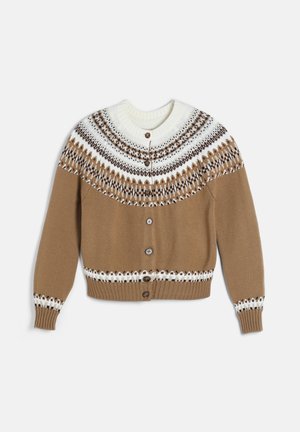 Femme portant un cardigan marron à motifs, une chemise blanche, un pantalon large en velours côtelé beige, une ceinture marron et des chaussures foncées, debout avec une main dans la poche.