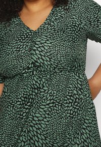 Femme portant une robe à col en V à motif feuillu vert et noir, manches courtes, taille élastique, une main posée sur la hanche.