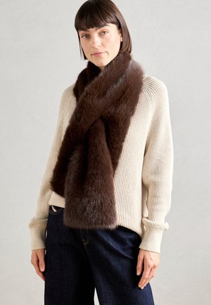 Kvinde iført beige strikket sweater, mørke bukser med vidde og et tykt mørkebrunt pels tørklæde draperet omkring halsen.