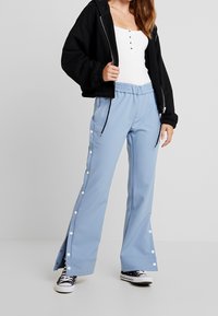 Femme portant un pantalon large bleu clair avec des boutons-pression sur les côtés, un débardeur blanc côtelé, une veste zippée noire et des baskets noires.
