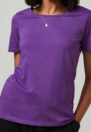 Femme portant un t-shirt violet uni à manches courtes et un collier en argent avec un petit pendentif rond, posant avec une main dans la poche d'un pantalon noir.