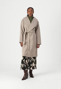Cappotto avvolgente in misto lana beige con un ampio colletto, vita con cintura e tasche laterali, indossato sopra una gonna a fantasia in bianco e nero e stivali al ginocchio.