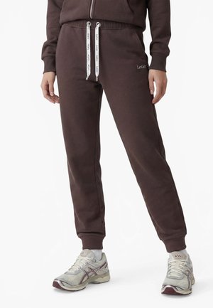 Personne portant un pantalon de jogging LeGer marron avec des cordons blancs et des baskets Asics blanches, debout, les mains détendues le long du corps.