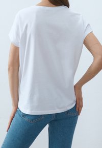 Bílé tričko s krátkým rukávem a kulatým výstřihem, hladká struktura a volný střih, v kombinaci s modrými džíny.