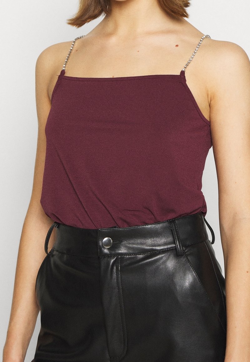 Personne portant un haut sans manches violet foncé avec de fines bretelles perlées et un pantalon en cuir noir taille haute avec fermeture par boutons.