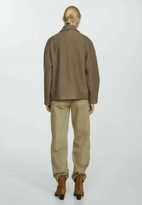 Brązowa kurtka z wełny z luźnym krojem, z detalami w postaci przeszyć i szerokim kołnierzem, zestawiona z jasnobeżowymi spodniami i brązowymi botkami na obcasie.