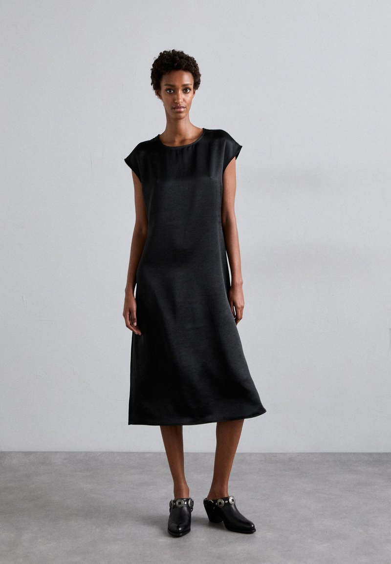 Black, short-sleeved dress with a loose fit, made from smooth fabric, falling to mid-calf, complemented by black clog shoes with metallic details.