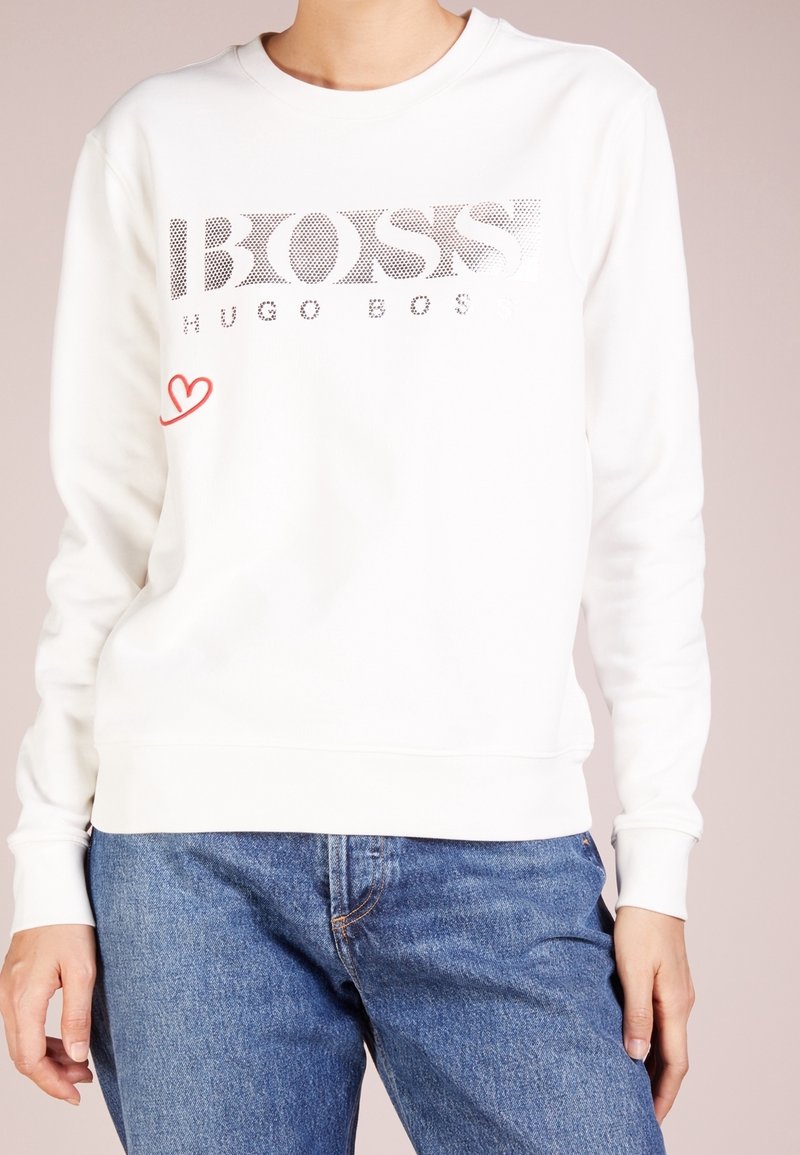 Personne portant un sweat-shirt blanc Hugo Boss avec un logo noir à pois et un petit détail de cœur rouge, associé à un jean en denim bleu.