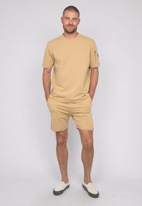 T-shirt beige et short assorti en tissu doux et léger. Comprend une poche sur la manche du t-shirt. Les chaussures sont des slip-ons écrus.