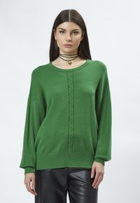 Grüner übergroßer Pullover mit gerippten Bündchen und Saum, der ein zentrales Zopfmuster-Detail aufweist. Kombiniert mit schwarzen Lederhosen.