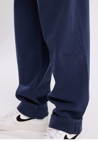 Navy blue trousers with a loose fit, cuffed hems, and a side pocket. Paired with white sneakers featuring a black logo.