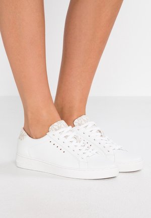Zapatillas blancas de corte bajo con lengüetas de talón beige con estampado, usadas descalzas sobre un fondo claro y liso.