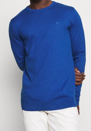 Blauer, langärmeliger Pullover aus Strickstoff, Rundhalsausschnitt, mit gerippten Bündchen und Saum. Kleines Logo-Detail auf der Brust. Kombiniert mit hellen Hosen.