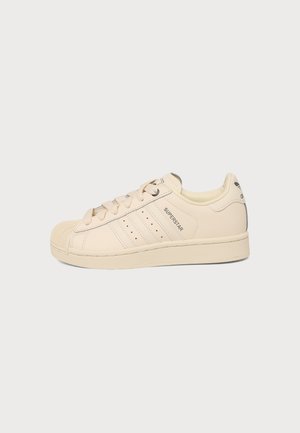 Baskets Adidas Superstar beige avec bout en forme de coquillage, trois bandes latérales, lacets et le texte "Superstar" sur fond blanc uni.