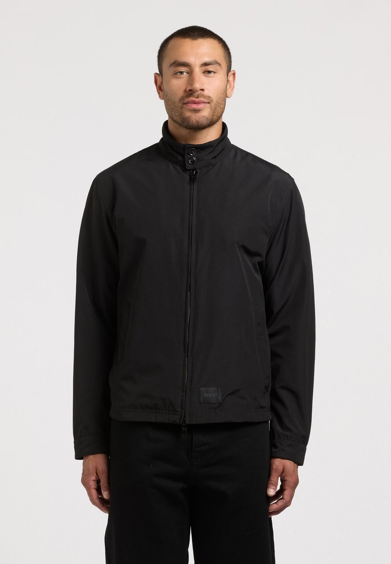 Homme debout face à l'avant, portant une veste noire à fermeture éclair avec un col montant et un pantalon noir, sur un fond uni.