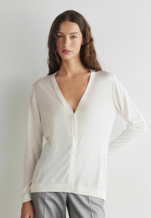 Frau mit langen braunen Haaren, die eine weiße, geknöpfte Strickjacke und graue Hosen trägt, steht mit einer Hand in der Hüfte vor weißem Hintergrund.