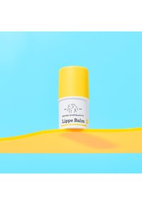 Drunk Elephant Lippe Balm in einem runden weißen Behälter mit einem gelben Deckel. Vor einem blauen Hintergrund mit einem gelben Wellenmuster präsentiert.