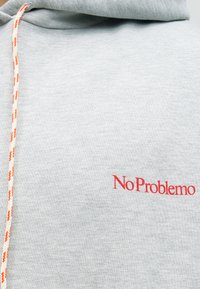 Ανοιχτό γκρι φούτερ με πορτοκαλί και λευκό κορδόνι και κόκκινο κέντημα με το κείμενο "No Problemo" στην περιοχή του στήθους.