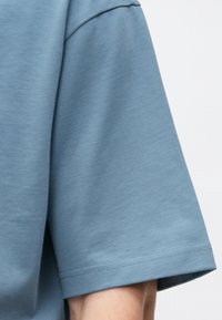 Brīvi pieguļoša, īsām piedurknēm zila krekla piedurkne uz personas, parādot auduma tekstūru un dubultā diega apdari.