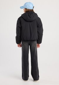 Black padded jacket with a hood, worn over loose-fitting gray jeans. A light blue cap completes the outfit. Subtle stitching details visible.